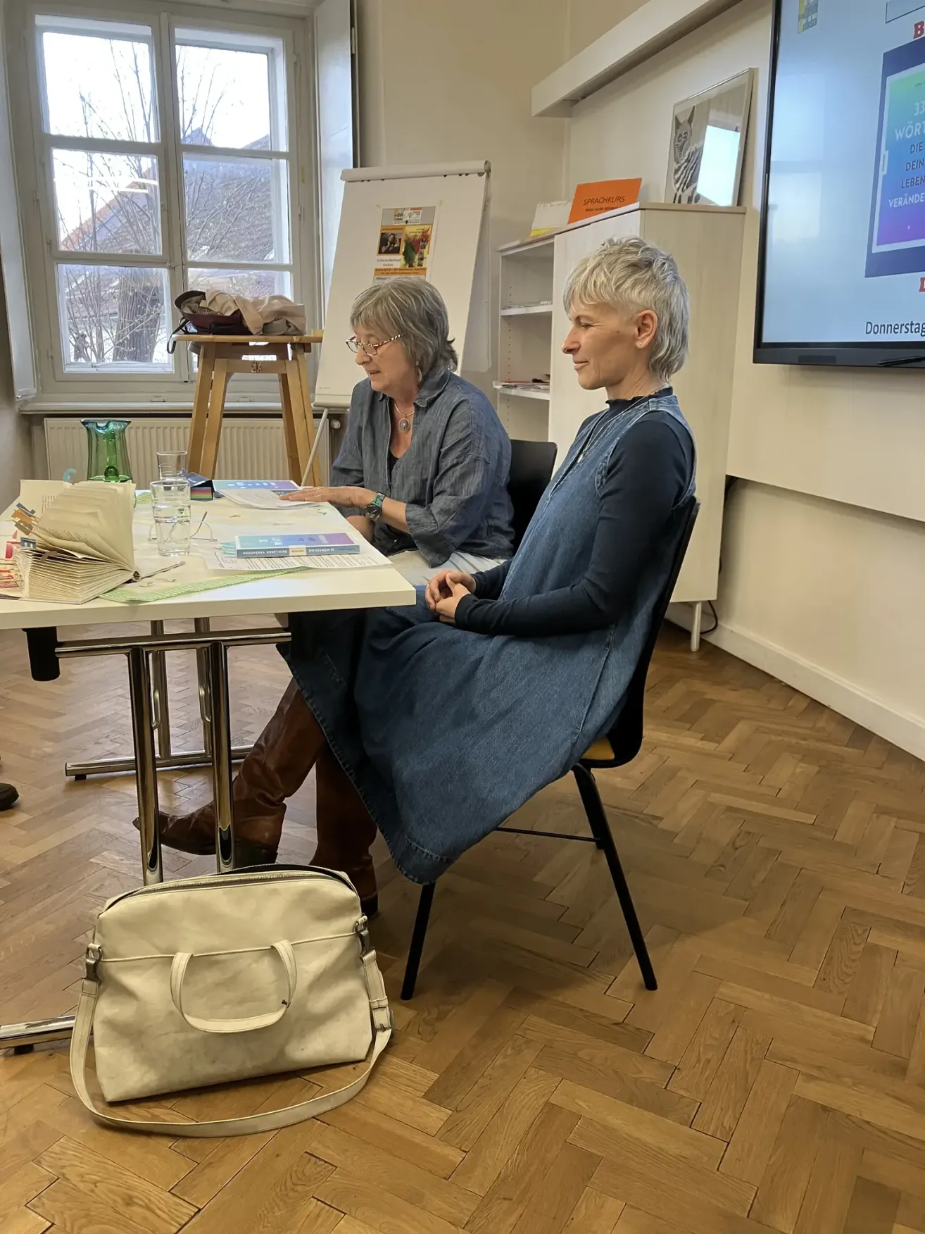 Two women sit at a table in a room with a large screen in the background. One is wearing glasses and a watch. The table has books, papers, and a glass. A handbag lies on the floor.