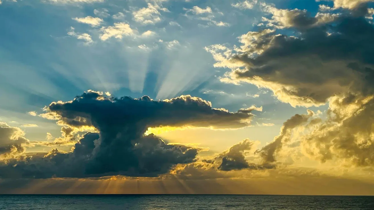A sunset scene with clouds casting rays of light over the ocean. The sky is blue with scattered clouds and the horizon is visible below.
