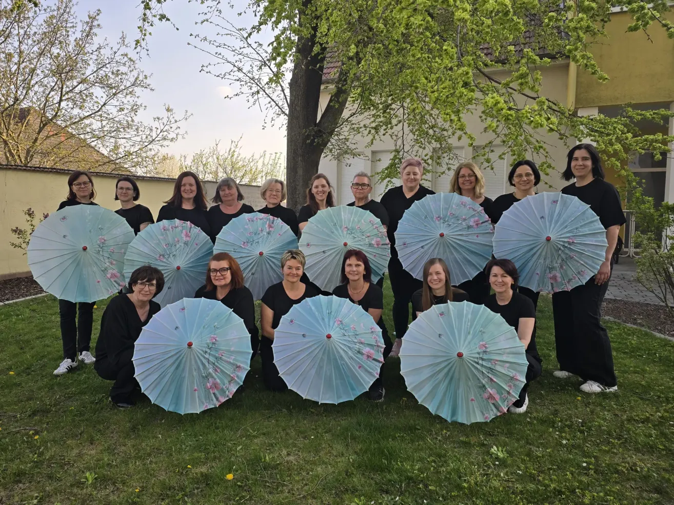 A group of women in black clothes are posing for a photo while holding decorative umbrellas with floral designs in front of a house with trees and a balcony.