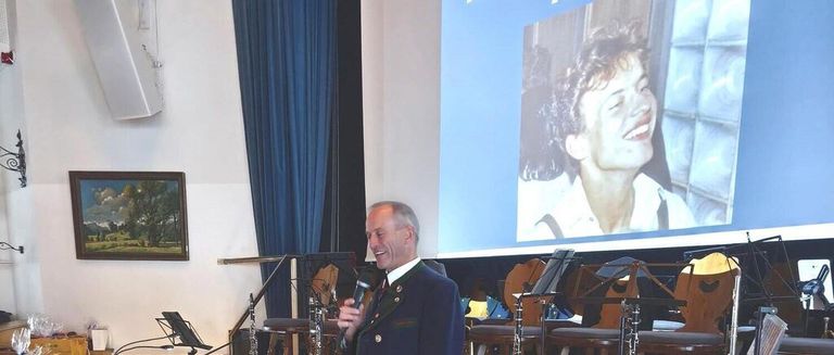 Ein Mann spricht in ein Mikrofon vor einer Projektion eines lächelnden Mannes und hält Klarinetten. Der Hintergrund besteht aus einer großen Leinwand und einer Landschaftsmalerei.