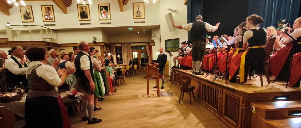 Eine Gruppe von Menschen in traditioneller Kleidung tritt auf einer Bühne auf, während das Publikum davor zusieht. Ein Mann leitet mit einem Mikrofon.