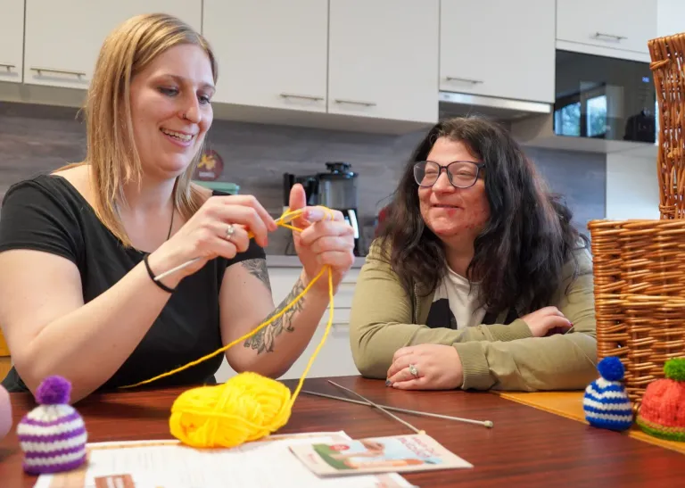 Zwei Frauen sitzen mit Stricknadeln an einem Tisch, eine mit einem Ball gelber Wolle, und lächeln sich an. Hinter ihnen eine Küche mit weißen Schränken und einem Herd.