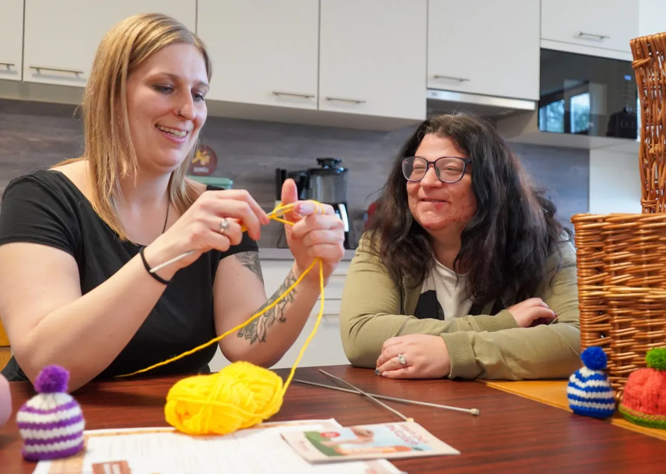 Zwei Frauen sitzen mit Stricknadeln an einem Tisch, eine mit einem Ball gelber Wolle, und lächeln sich an. Hinter ihnen eine Küche mit weißen Schränken und einem Herd.