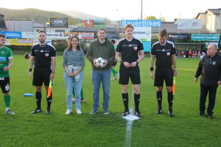Five people stand on a soccer field, two men holding a soccer ball, and three men in black uniforms. Behind them, a woman in blue jeans stands and smiles. A white line and flags are on the ground.
