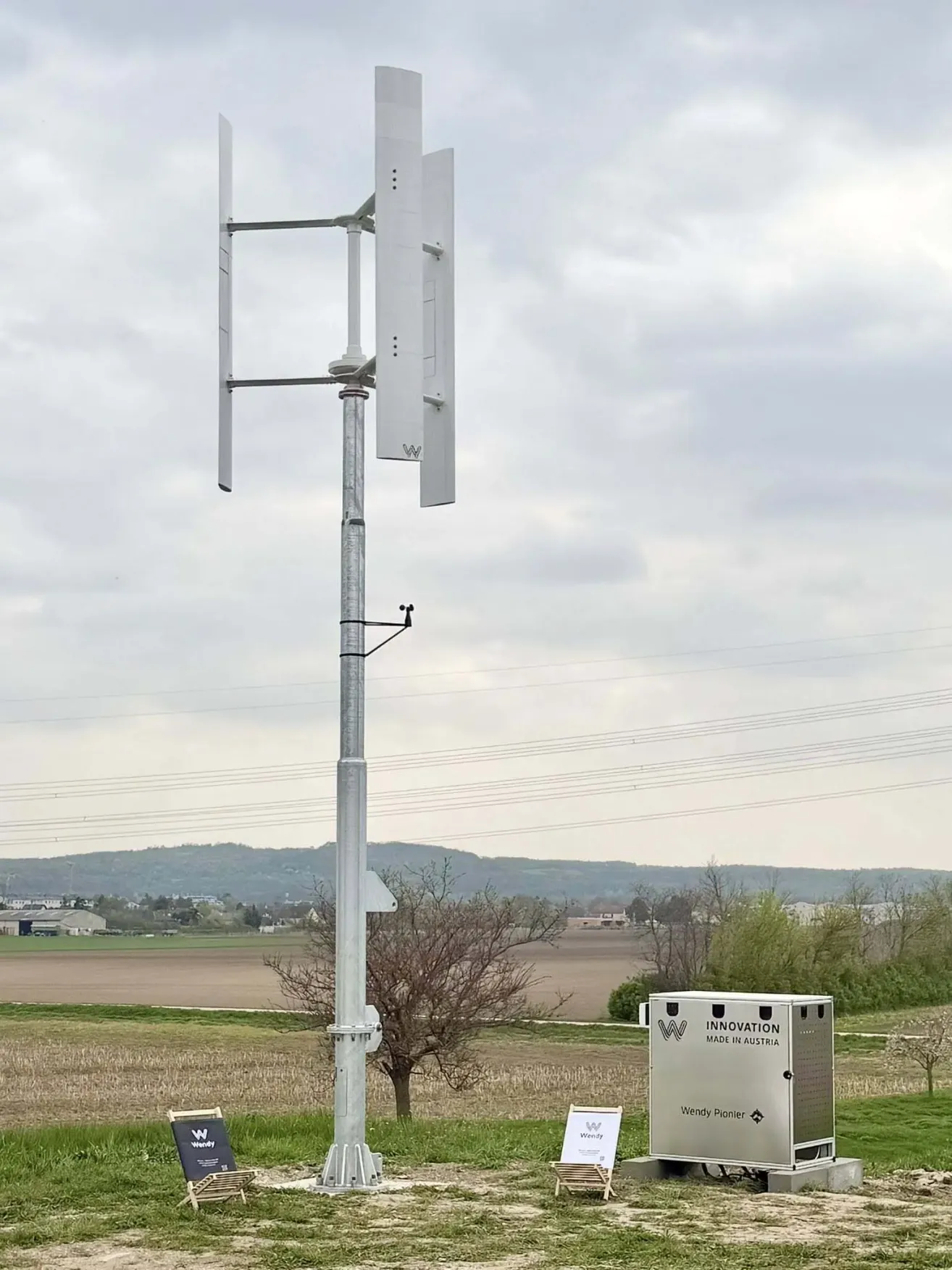 Eine vertikale Windturbine steht auf einer Wiese bei klarem Himmel und Hügeln in der Ferne. In der Nähe ist ein Kasten mit dem Wort 'Innovation' zu sehen.