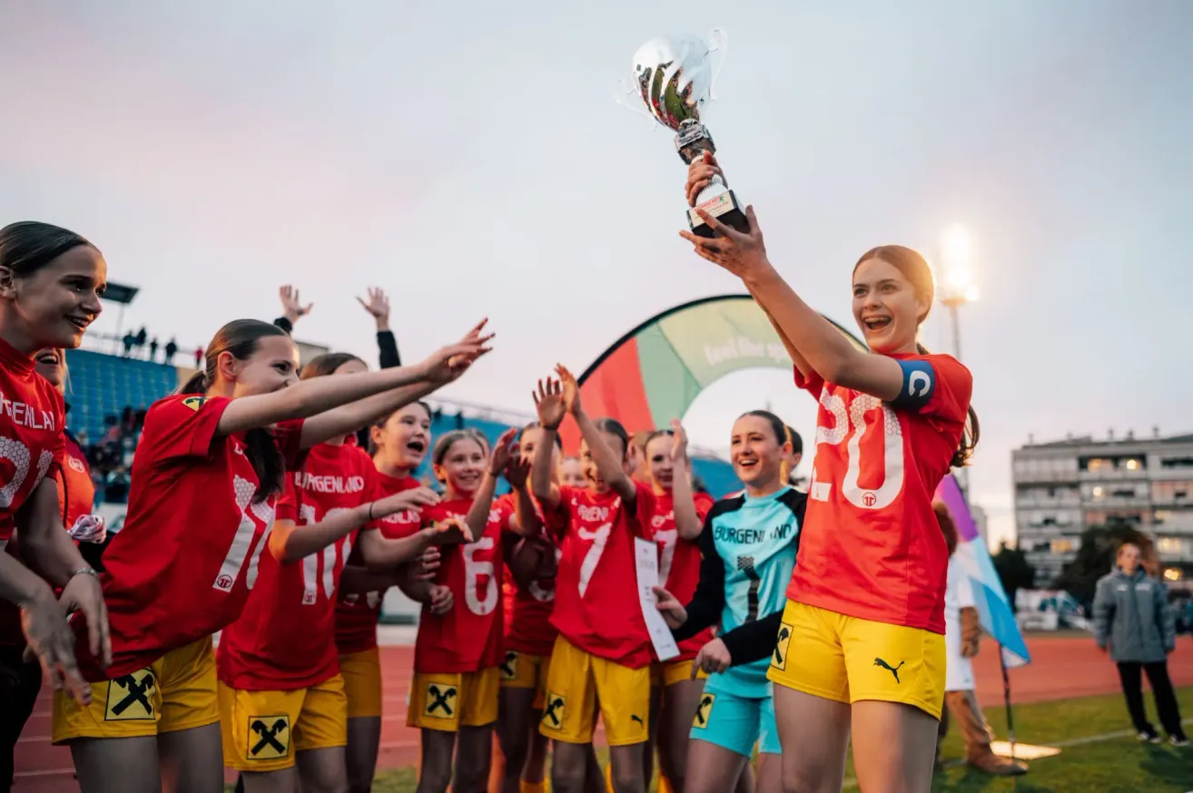 A team of young women wearing red and yellow uniforms, celebrating with a trophy on a field at dusk.