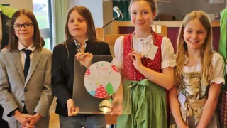 Three girls hold a glass plaque with a Christmas design and the words 'KiGra PREMSTATTEN'. The girl in the middle wears a traditional dress. Behind them, a microphone on a stand is visible.