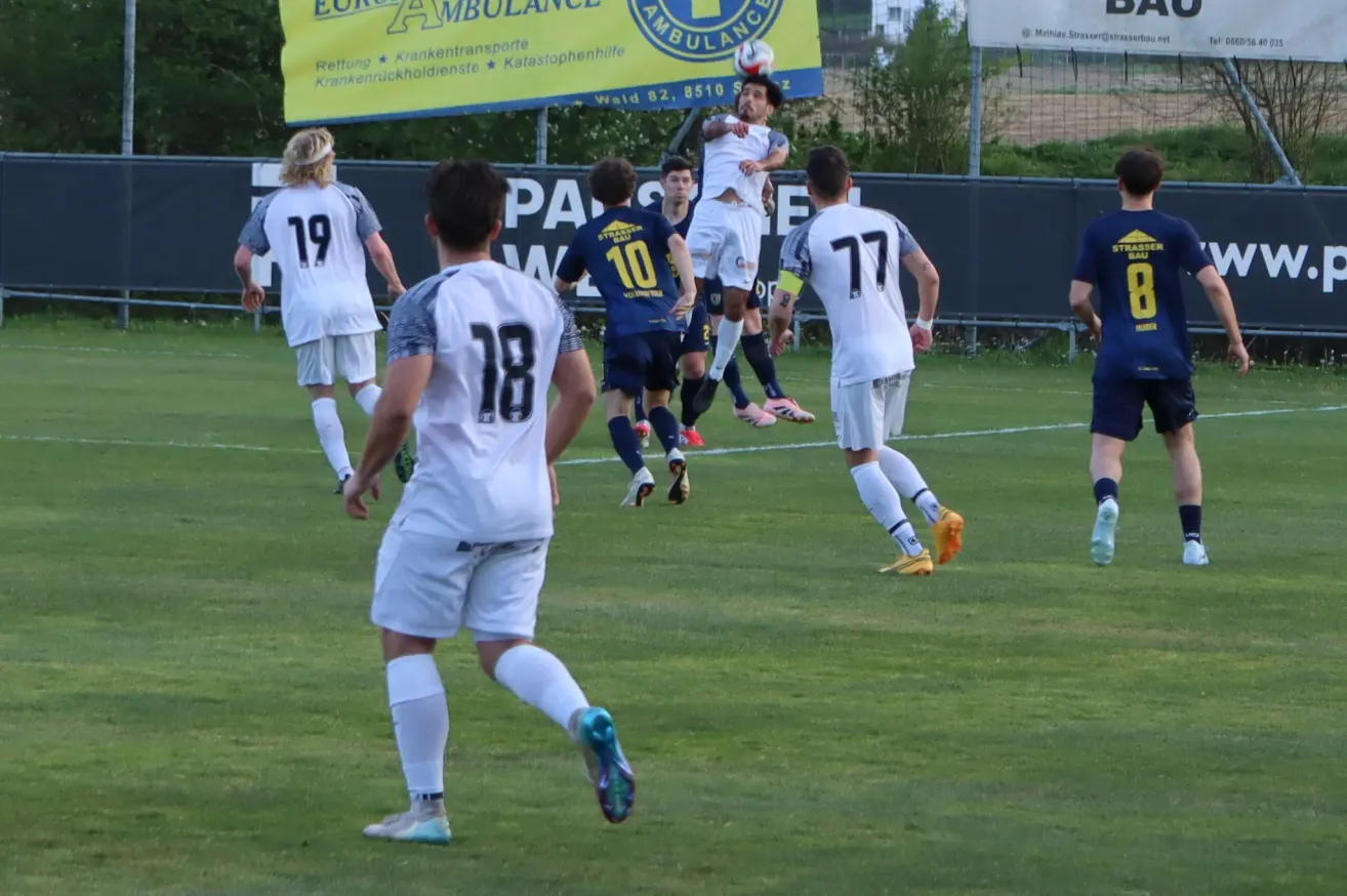 A soccer match with players in white and blue uniforms, number 18 jumps to head the ball. The background shows a banner with 'Ambulance' and a soccer ball in the air.