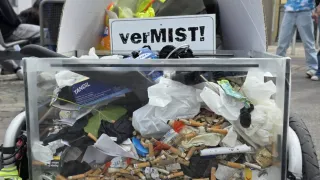 A glass container filled with trash, cigarette butts, and other garbage is mounted on a cart. A sign with the word 'verMIST!' is attached. A man and a girl are standing behind the cart.