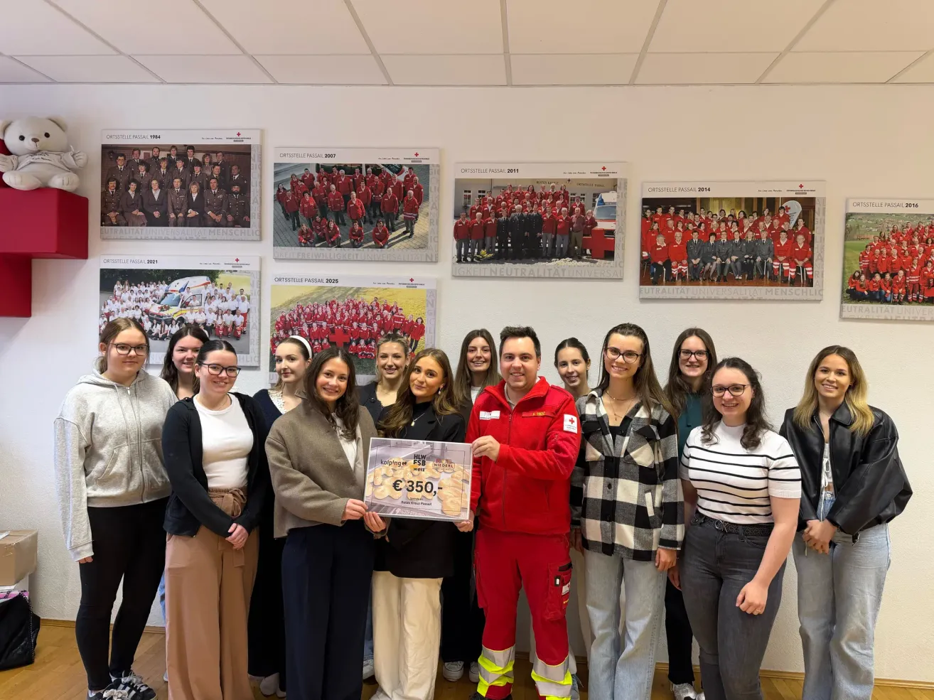 A group of smiling people stand around a man in red, holding a certificate for 350 euros. Behind them, framed photos of groups in red uniforms adorn the wall.