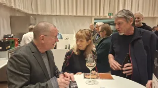 A man signs a book at a table while two women and another man look on. A white table holds wine glasses and brochures. Curtains hang in the background.