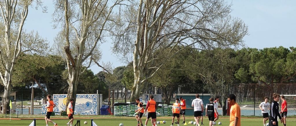 Eine Gruppe junger Athleten trainiert auf einem von Bäumen und einem Zaun umgebenen Fußballfeld. Das Feld hat Gras und Fußballbälle. Die Spieler tragen orange Weste und Turnschuhe.