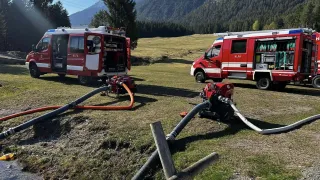 Two red fire trucks are parked in a grassy field with mountains in the background. One has its back door open, and both have hoses connected to water sources.