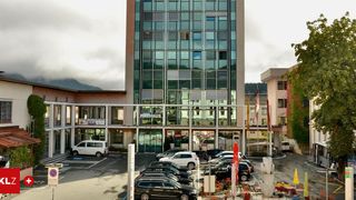 A large building with glass windows has parked cars in front. The building has the text Rathaus on it. There are flags on poles and plants on the side. A white van is parked on the left. A white building is on the right side.