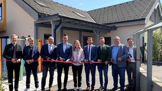 Eine Gruppe von Männern und Frauen in Geschäftskleidung steht vor einem Gebäude. Sie halten eine Schleife zum Schneiden.