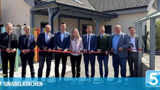 Eine Gruppe von Menschen in formeller Kleidung steht vor einem Gebäude und schneidet ein rotes Band ab.