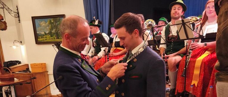 Ein Mann heft eine Medaille an die Jacke eines anderen Mannes, der lächelt. Ein Musiker-Band sitzt hinter ihnen, einer spielt Trompete.