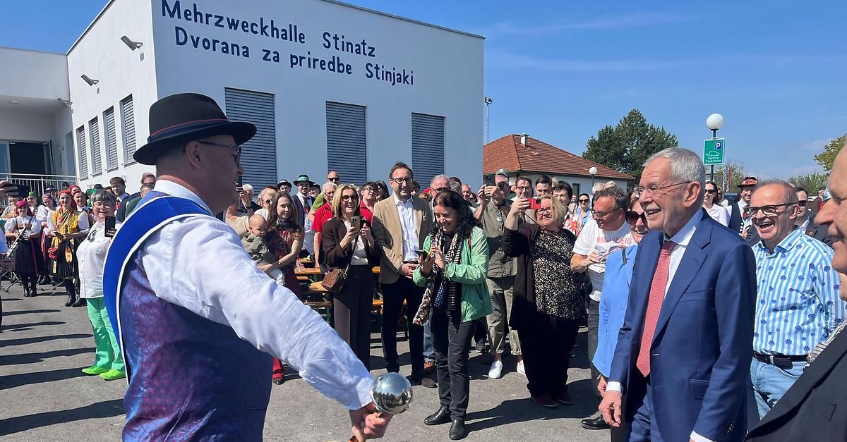 A crowd gathers outside a building with 'Mehrzweckhalle Dvorana za priredbe Stinjaki' on the wall. A man in a hat juggles a silver ball. Some people take photos.