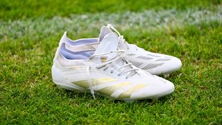 Ein Paar weißer Fußballschuhe mit goldenen Akzenten liegt auf einem Grasfeld. Die Schuhe haben ein schwarz-weißes Muster.