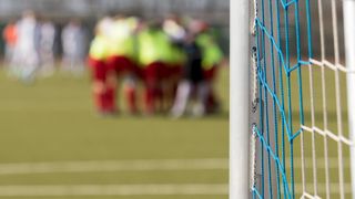 A blurry view of a soccer team in green and red uniforms, standing close together, possibly strategizing. The goal net is blue.