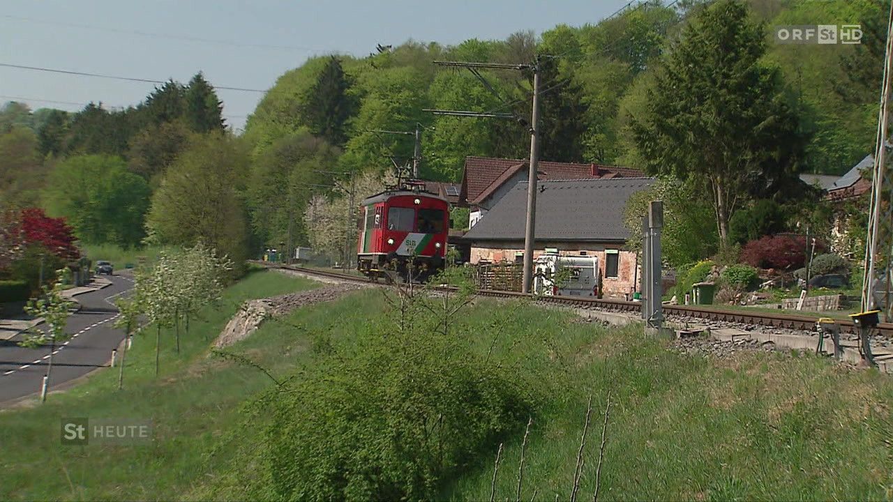 A red train is on the track with a house in the background. The train has a green roof. Trees surround the area.