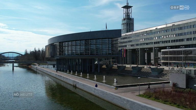 Ein modernes Gebäude mit Glasfenstern und Flaggen steht neben einer Wasserfontäne, an der mehrere Menschen vorbeigehen.