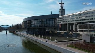 Ein modernes Gebäude mit Glasfenstern und Flaggen steht neben einer Wasserfontäne, an der mehrere Menschen vorbeigehen.