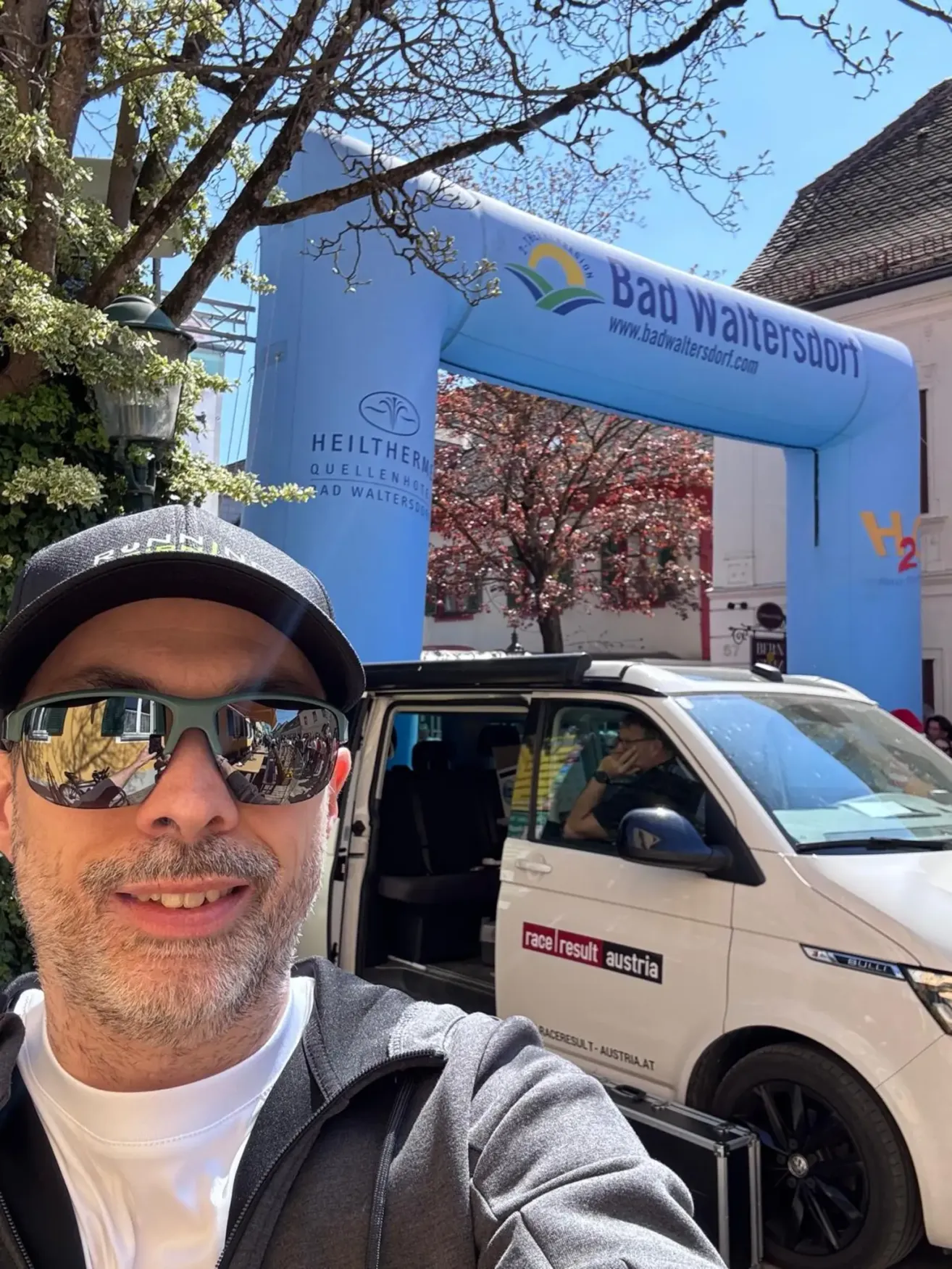 A man wearing sunglasses and a cap takes a selfie in front of a van, with a blue arch reading 'Bad Waltersdorf' in the background.