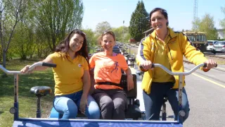 Three women in yellow shirts sit on a tricycle. The woman in the middle wears a T-shirt with the year 2023 and a logo. Behind them are trees and a tower.