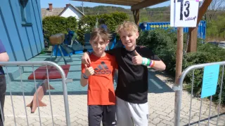 Two boys stand in front of a numbered sign, one with an orange shirt and the other with a black shirt. Behind them is a playground.