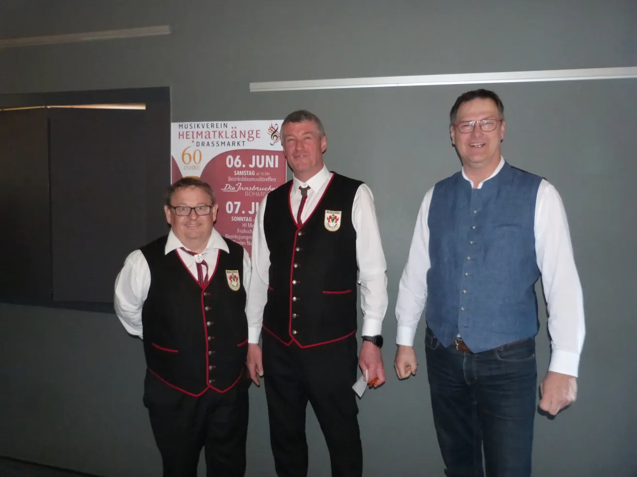 Three men in traditional clothing stand next to a poster. The man on the left wears a black vest with red trim, the middle man a black vest with red and white trim, and the man on the right a blue vest. They are smiling and appear to be at an event.