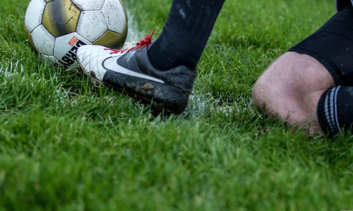 A soccer player wearing Nike cleats kicks a ball on a grassy field, showing mud and splashes.