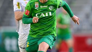 Zwei Fußballspieler auf dem Feld, einer in Grün jagt dem Ball hinterher, der andere in Weiß versucht, ihn zu blockieren.