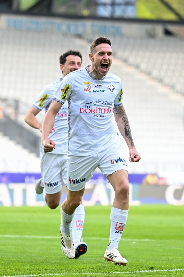 Zwei Fußballspieler in weißen Trikots feiern ein Tor auf einem Feld. Ein Spieler läuft mit offenem Mund, während der andere hinter ihm lächelt. Leere Tribünen im Hintergrund.