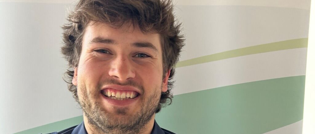 Ein Mann mit Bart und kurzen, lockigen Haaren lächelt und posiert für ein Foto. Er steht vor einer weißen Wand mit grünen Streifen.