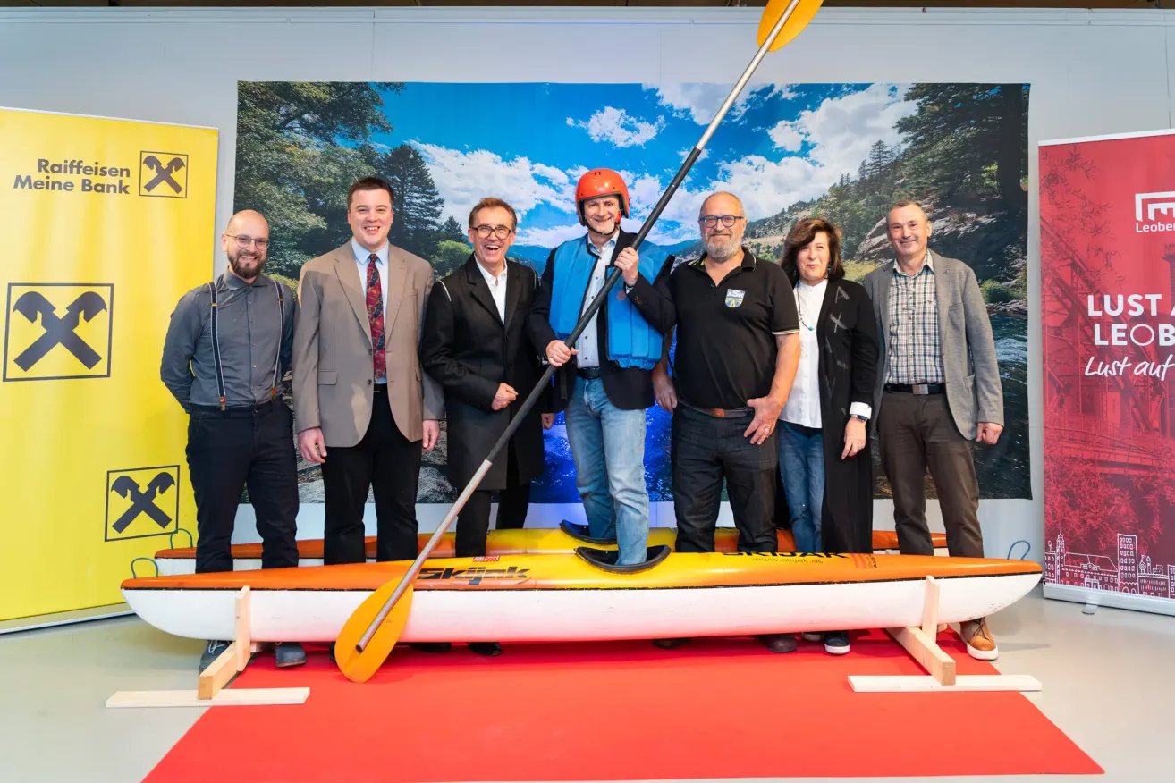 Eine Gruppe von Menschen steht hinter einem Kajak und lächelt für ein Foto. Ein Mann hält eine Paddel. Ein malerischer Hintergrund ist hinter ihnen.