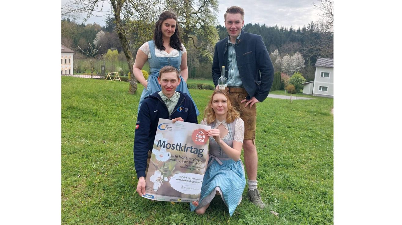 Vier Personen posieren für ein Foto auf einem Rasen mit Bäumen und einem Berg im Hintergrund. Zwei Männer und zwei Frauen. Ein Mann hält ein Plakat, ein anderer eine Flasche. Die Frauen tragen traditionelle Kleider.