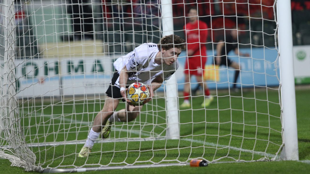 Ein Fußballspieler in einem weißen Trikot und schwarzen Shorts taucht, um einen Ball zu blocken, seine Augen auf den Ball gerichtet, während zwei andere Spieler in roten und schwarzen Trikots im Hintergrund zusehen.