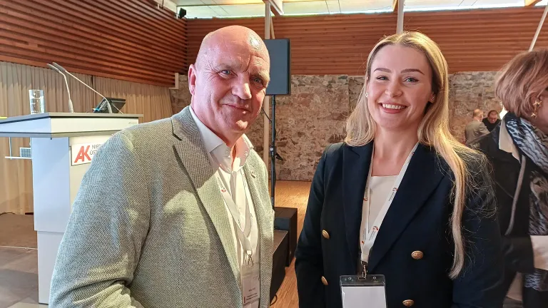 A man and a woman are smiling for a photo in an event hall. The man is wearing a gray suit and the woman a navy blue blazer. Both have white ID cards on lanyards. Behind them is a speaker and a stone wall.