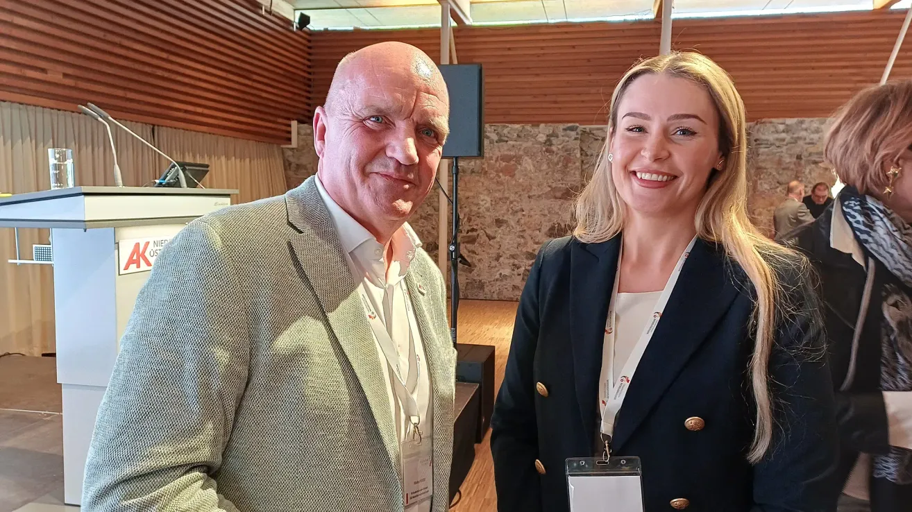 A man and a woman are smiling for a photo in an event hall. The man is wearing a gray suit and the woman a navy blue blazer. Both have white ID cards on lanyards. Behind them is a speaker and a stone wall.