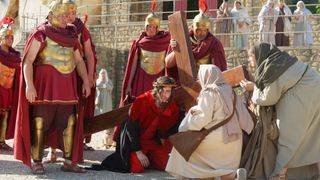 A man wearing a crown of thorns kneels as soldiers hold a wooden cross. Behind him, a woman offers support. Other soldiers and onlookers are present.