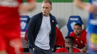 A man in a blue jacket and white shirt walks down the sideline of a sports field with others sitting in the background.