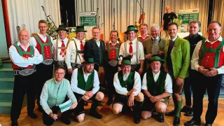 Eine Gruppe von Männern in traditioneller deutscher Kleidung posiert für ein Foto, mit einem Hintergrund, der ein Banner mit deutschem Text zeigt.