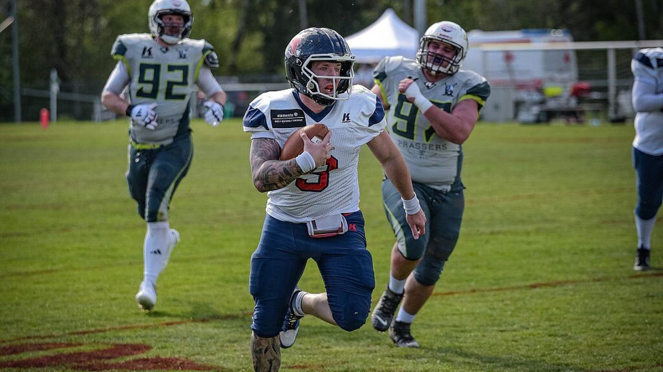 Three American football players in helmets run on the field. The central player holds a ball. The other two players follow him. Behind them are trees and a white tent.