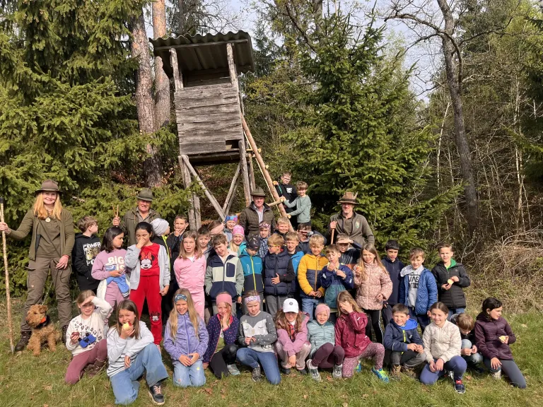 Eine große Gruppe von Kindern und Erwachsenen posiert für ein Foto im Wald. Hinter ihnen steht ein Baumhaus. Die Kinder tragen Winterkleidung.