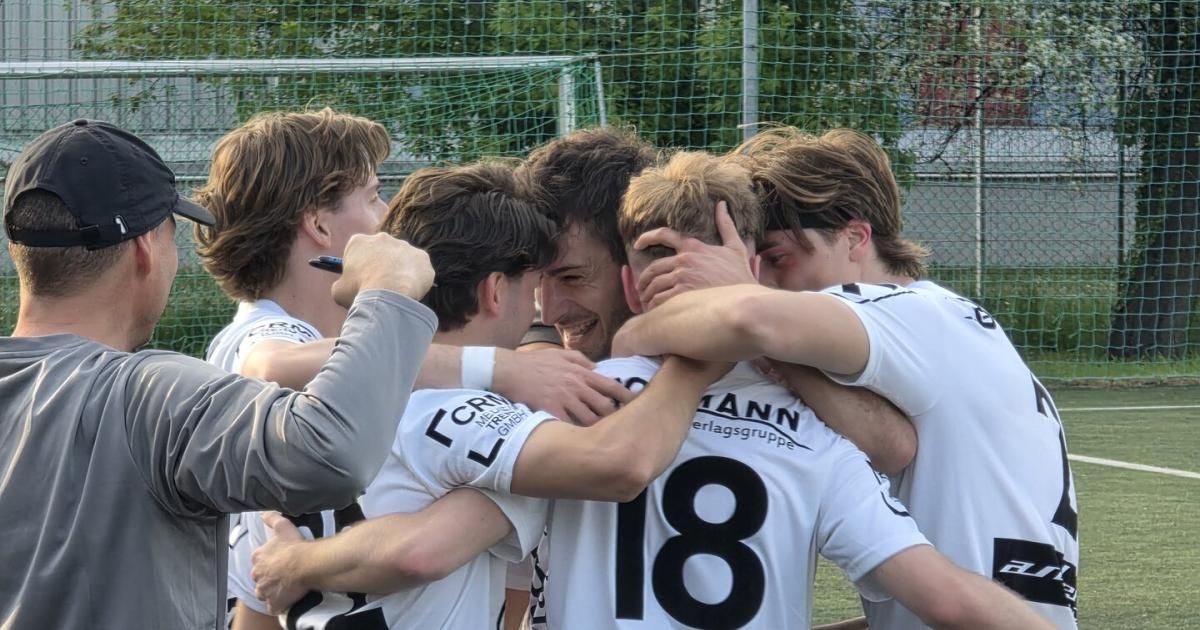 Eine Fußballmannschaft in weißen Trikots mit Zahlen und Buchstaben schmiegt sich vor einem Tor zusammen, ein Mann in Grau hält einen Stift.