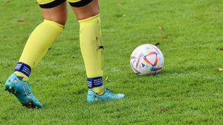 A soccer player in yellow socks and blue shoes stands on a field with a colorful soccer ball. The player's socks are muddy.