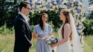 Eine Hochzeitszeremonie mit einem Mann in einem Anzug, einer Frau in einem blauen Kleid und einer Braut in einem weißen Kleid, alles unter einem Blumenbogen.