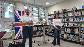 Two individuals are standing in a library with a British flag. A man is wearing glasses and holding a book. A woman is wearing a white shirt and standing with her hands clasped.