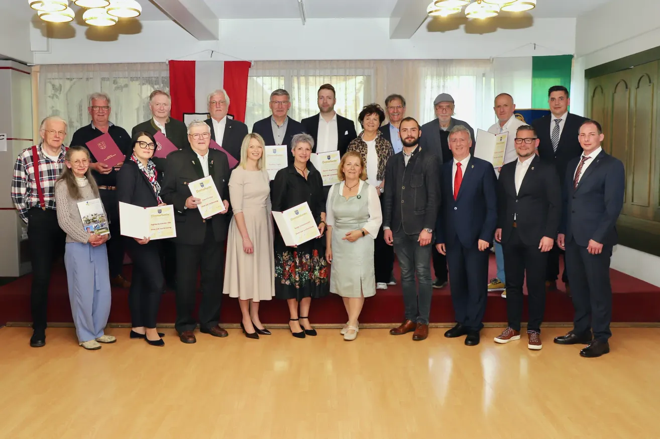 A group of people are posing for a photo at an awards ceremony. They are holding certificates and standing on a stage. There are red banners on the wall behind them.